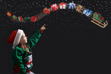 Cute little girl in Santa hat pointing at gift boxes flying out of sledge on black background. Christmas celebrationの写真素材