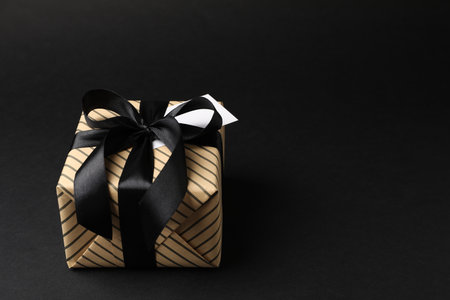 Beautiful wrapped gift box with card on black background, closeup. Space for textの写真素材