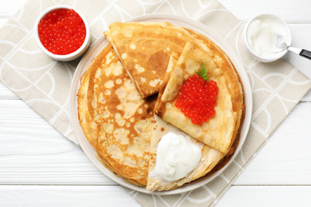 Tasty crepes with red caviar and sour cream on white wooden table, flat lay