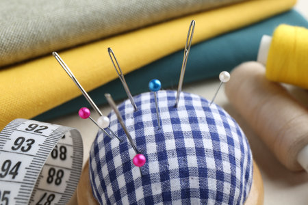Checkered pincushion with pins, needles, spools of threads, fabrics and measuring tape on light...
