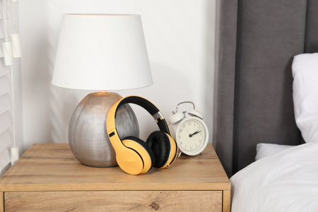 Stylish headphones, lamp and alarm clock on bedside table in bedroom, closeup