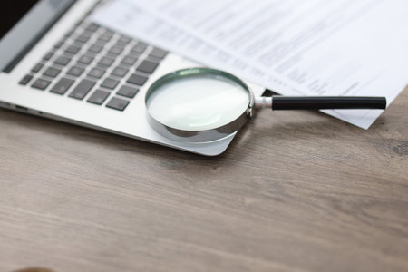 Magnifying glass, laptop and documents on wooden table, closeup. Space for textの写真素材
