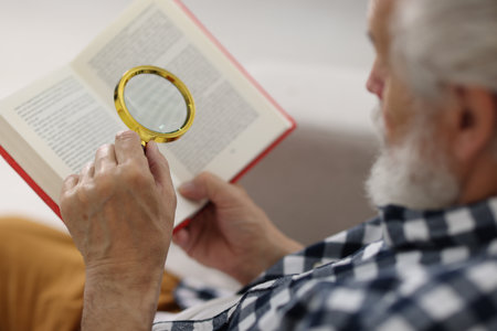 Senior man using magnifying glass while reading book indoors, closeupの写真素材