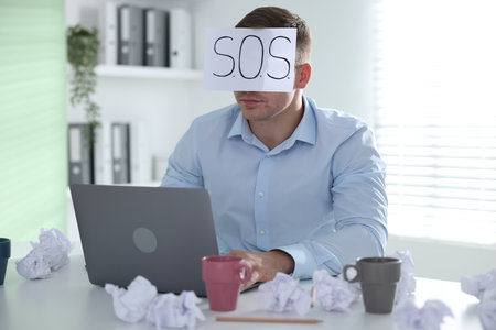 Man with laptop and sign SOS at table in office