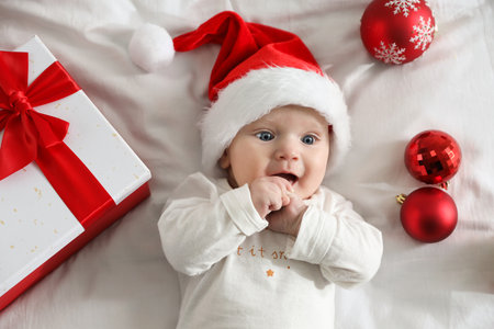 Baby in Santa hat, Christmas gift and baubles on bed, top view