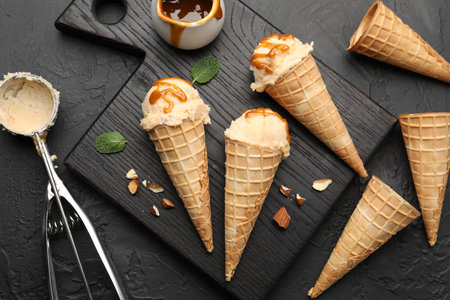 Tasty ice cream with caramel sauce in waffle cones on black table, flat lay