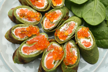Tasty spinach crepes with salmon, cream cheese and leaves on plate, top view