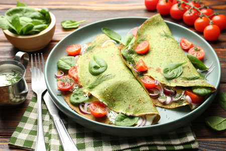 Tasty spinach crepes with tomatoes, onion, sour cream and leaves on wooden table, closeupの写真素材