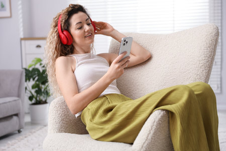 Happy woman in headphones with smartphone listening music on armchair at homeの写真素材