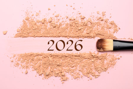 Makeup brush, crushed decorative cosmetic product and numbers 2026 on pink background, top view