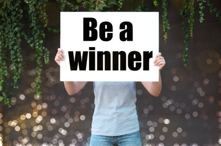 Woman holding poster with text Be A Winner outdoors with bokeh lights, closeupの写真素材