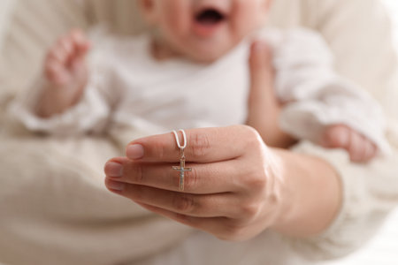Sacrament of baptism. Woman holding little baby and gold cross indoors, selective focus
