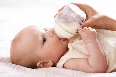 Mother feeding her newborn baby with bottle on bed indoors, closeup