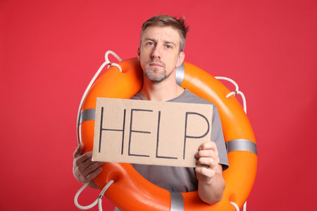 Unhappy man with lifebuoy and cardboard sign with word Help on red backgroundの写真素材