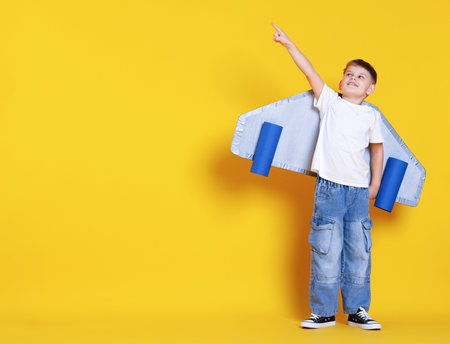 Little boy with cardboard plane wings pointing at something on yellow background. Space for textの写真素材
