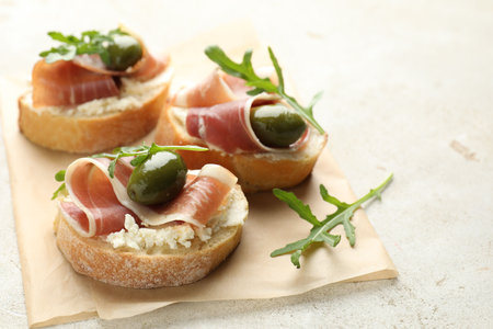 Delicious bruschettas with cream cheese, prosciutto, green olives and arugula on gray textured table, closeup. Space for textの写真素材