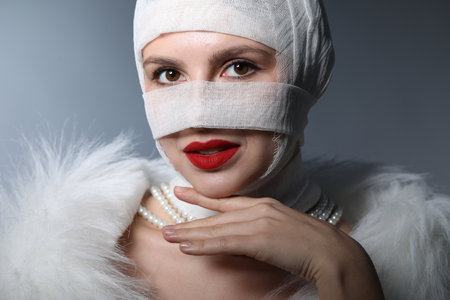 Woman with face wrapped in medical bandage after plastic surgery operation on gray backgroundの写真素材