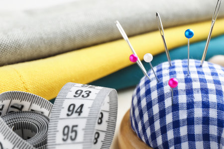 Checkered pincushion with pins, needles, fabrics and measuring tape on light table, closeup