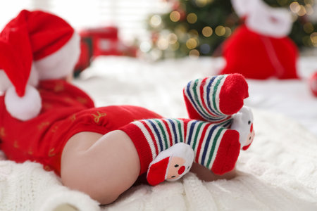 Cute baby in Santa hat and Christmas socks on bed against blurred lights indoors, selective focus