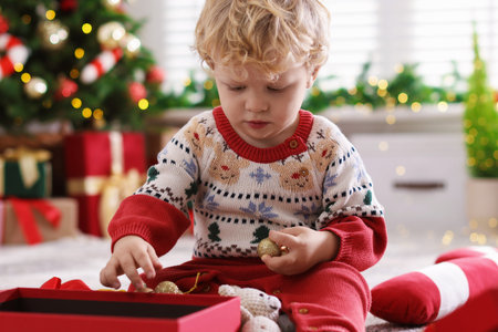 Cute little baby with Christmas ball and gift box on floor in festive decorated roomの写真素材