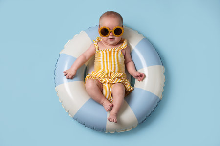 Cute little baby with sunglasses on inflatable ring against light blue background, top view