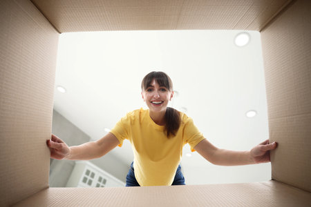 Woman looking into cardboard box against ceiling in room, view from inside package