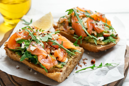 Tasty bruschetta with salmon, avocado, arugula, seeds and feta cheese on table, closeup