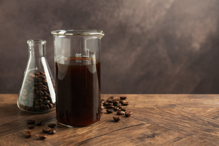 Glass beaker with coffee and beans in flask on wooden table against brown background, closeup. Space...