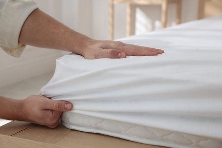 Man putting bed sheet on mattress indoors, closeup