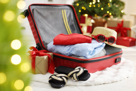 Suitcase with clothes, sunglasses, shoes and gifts on floor in festive decorated room, closeup....