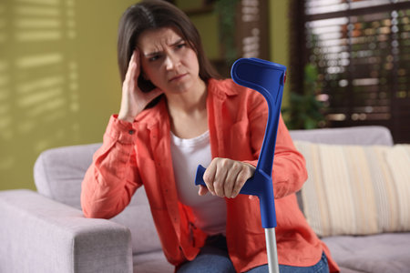 Unhappy woman with elbow crutch sitting on sofa at home, selective focus