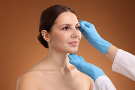 Doctor checking patient's face before plastic surgery operation on brown gradient background,...
