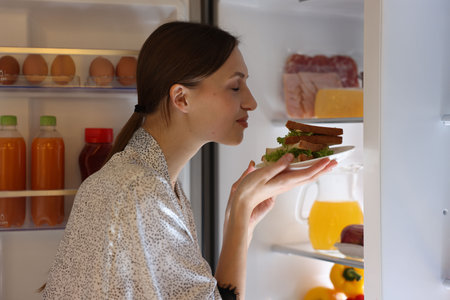 Woman with tasty sandwiches near open refrigerator indoors
