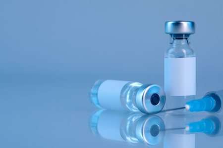 Vaccine in glass vials and syringe on mirror surface against gray background, closeup. Space for...