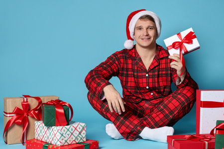 Happy man in Santa hat and pajamas with Christmas gifts on light blue backgroundの写真素材