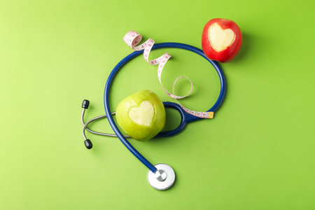 Ripe apples with carved hearts, measuring tape and stethoscope on green background, flat layの写真素材