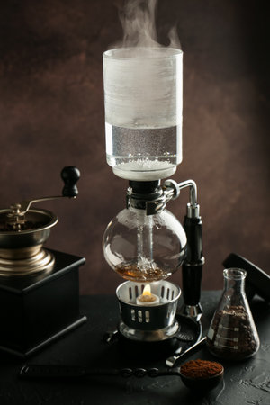 Siphon (vacuum) coffeemaker with hot water and coffee grinder on black table against brown background, closeupの写真素材