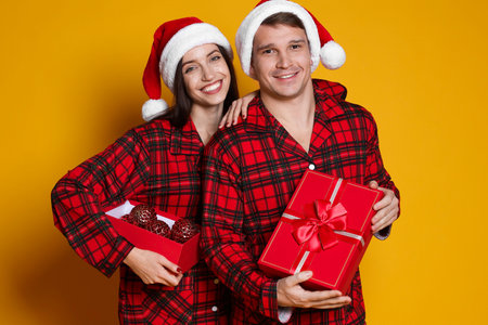 Happy couple in Santa hats and pajamas with Christmas balls on orange backgroundの写真素材