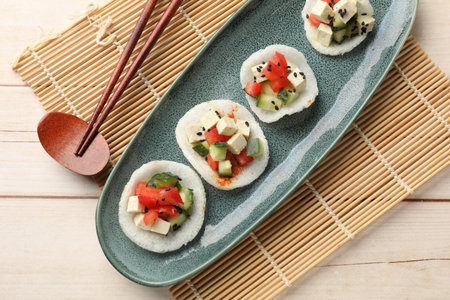 Tasty rice paper chips with vegetables, feta cheese and black sesame seeds on light wooden table,...