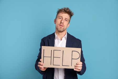 Unhappy businessman holding cardboard sign with word Help on light blue background
