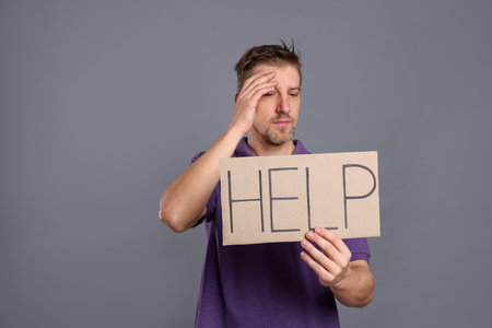 Unhappy man holding cardboard sign with word Help on grey background