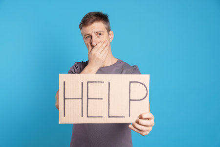Unhappy man holding cardboard sign with word Help on light blue background