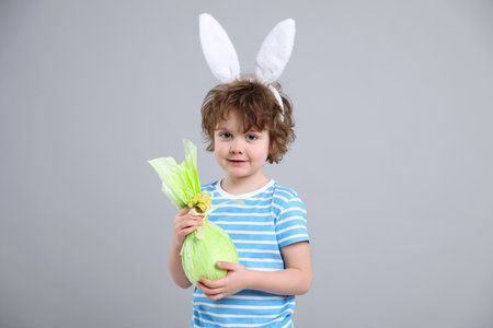 Easter celebration. Portrait of cute boy in headband with bunny ears holding wrapped gift on grey...