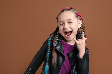 Cute girl with stylish braids showing rock gesture on brown background