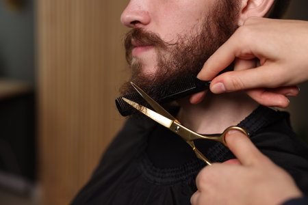 Barber trimming man's beard in barbershop, closeup. Space for text