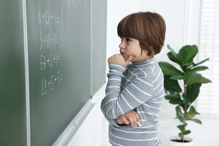 Thoughtful schoolboy near chalkboard with math assignment in classroom