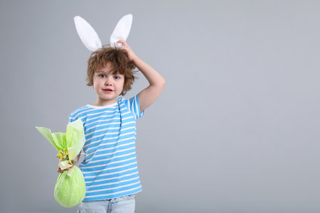 Easter celebration. Portrait of cute boy in headband with bunny ears holding wrapped gift on grey...