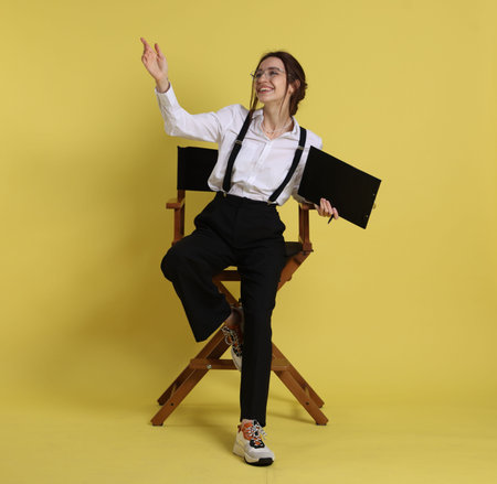 Professional film director with clipboard pointing at something on chair against yellow background