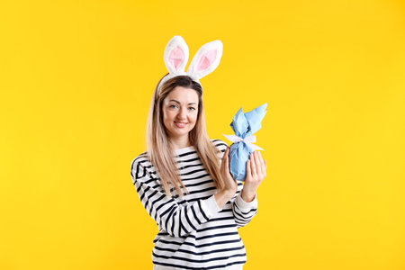 Easter celebration. Smiling woman in headband with bunny ears holding wrapped gift on yellow...