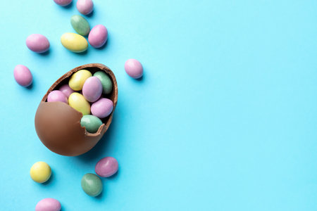 Easter celebration. Broken chocolate egg with candies on light blue background, flat lay. Space for...